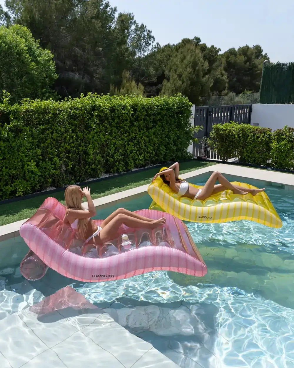 Duas amigas a relaxar lado a lado em colchões insufláveis rosa e amarelo na piscina